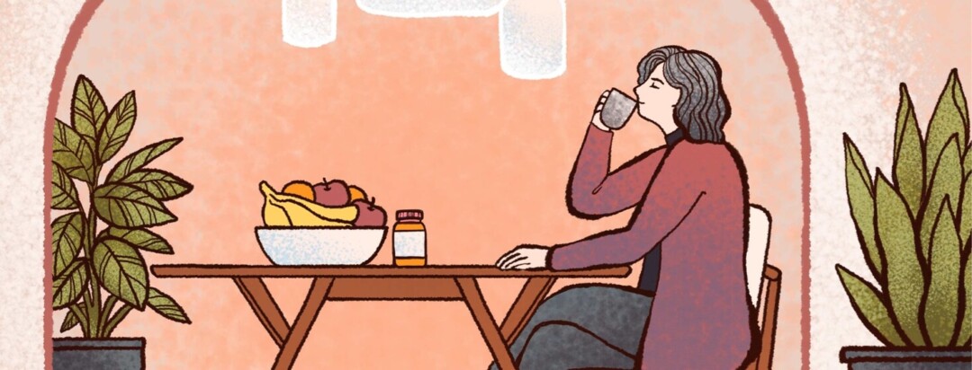 A woman sits at a table with fruit and vitamins as she sips calmly from a mug.