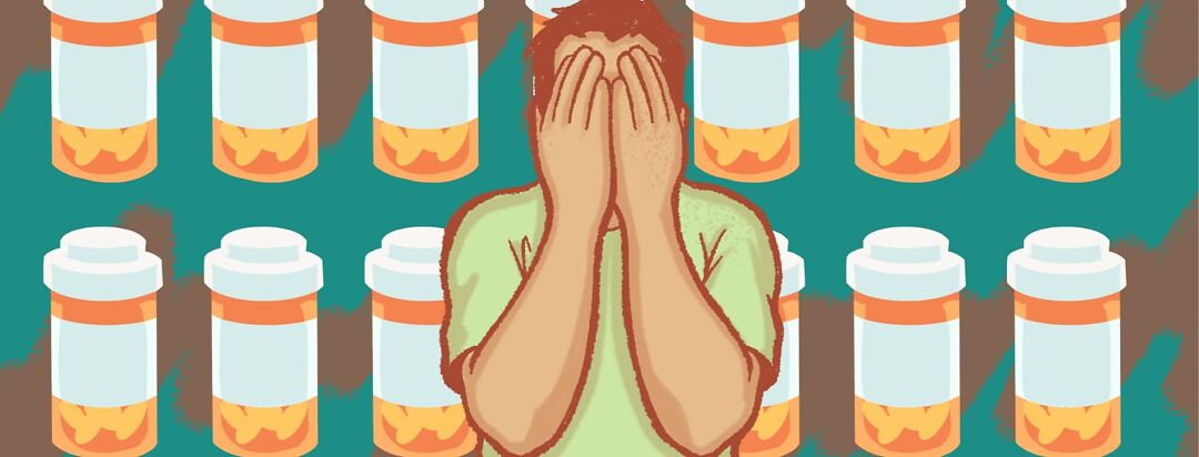 A man is overwhelmed with his hands covering his face while medications pile up behind him.